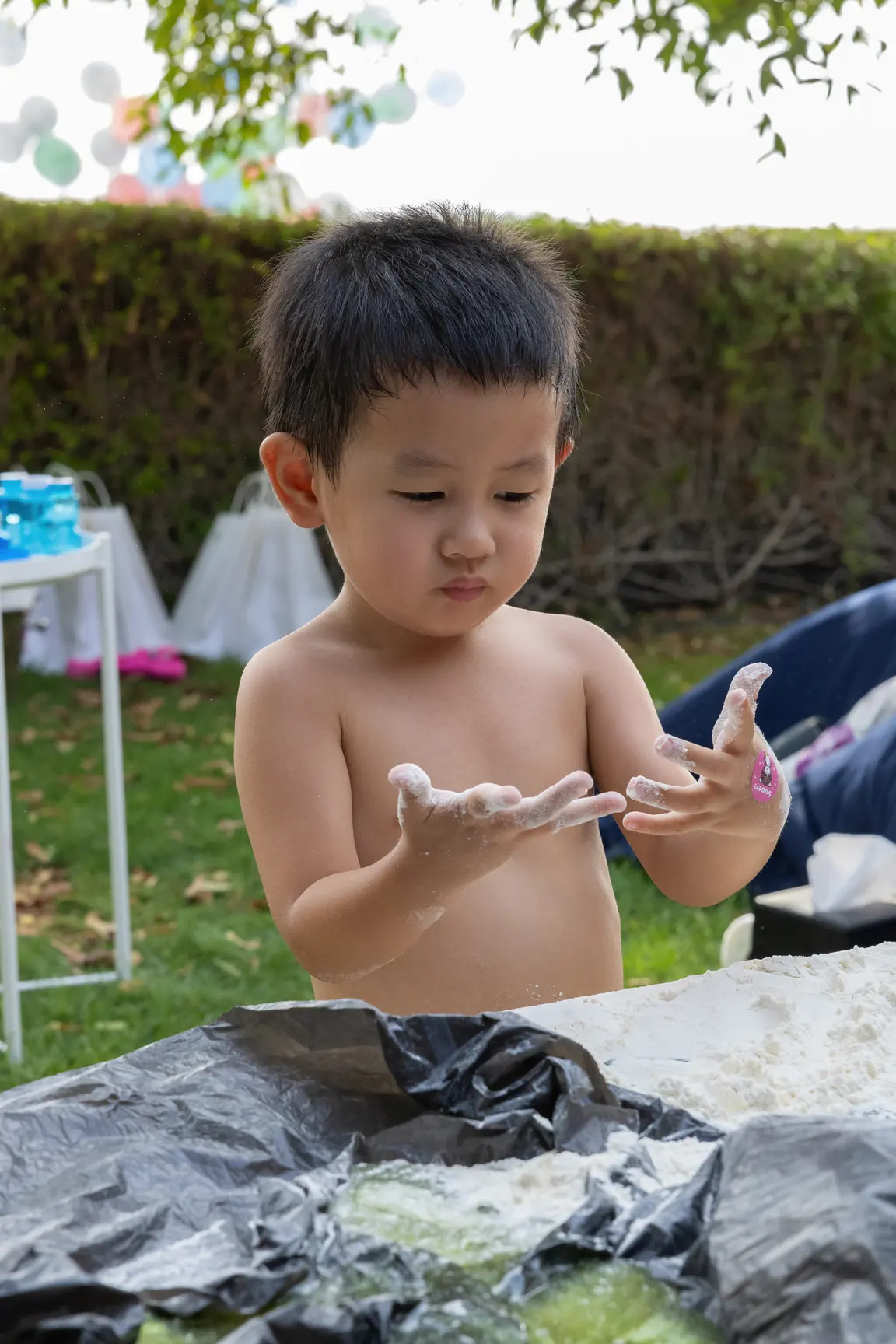 Young child, shirtless, outdoors, hands covered in white powder or flour, concentrating on their fingers during a activity at a table.