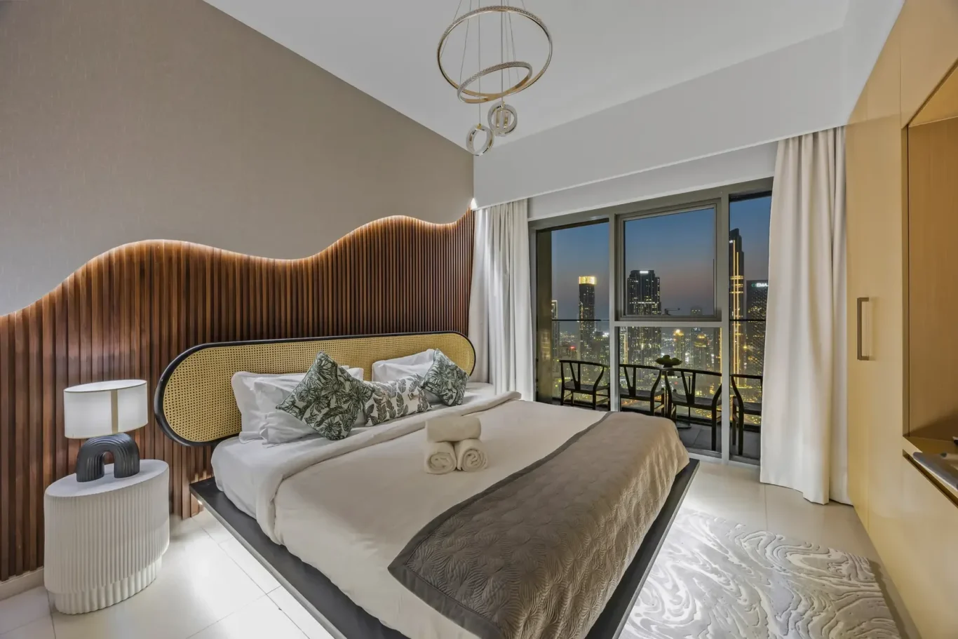 Luxurious hotel bedroom with a wavy wood headboard, neutral bed linens, and a city skyline visible through large windows at dusk.
