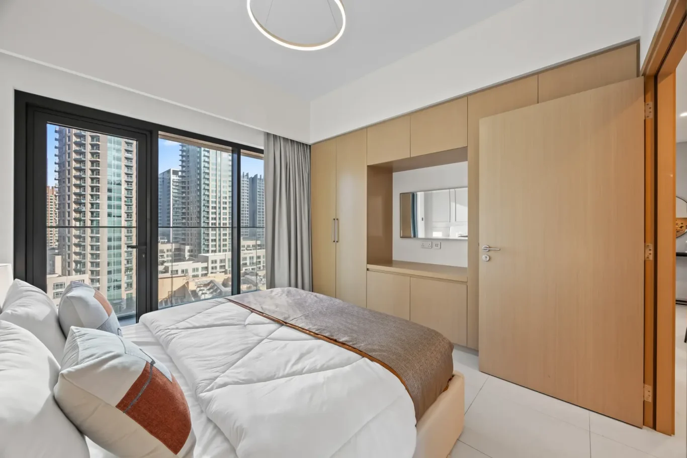 Twin beds in a bright modern hotel room, with a large window showing a city skyline and built-in light wood wardrobes.
