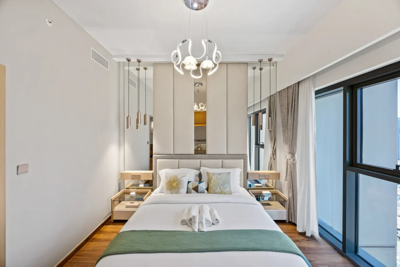 Modern hotel bedroom with a padded headboard, two gold bedside tables, pendant lights, and a large window with drapes.