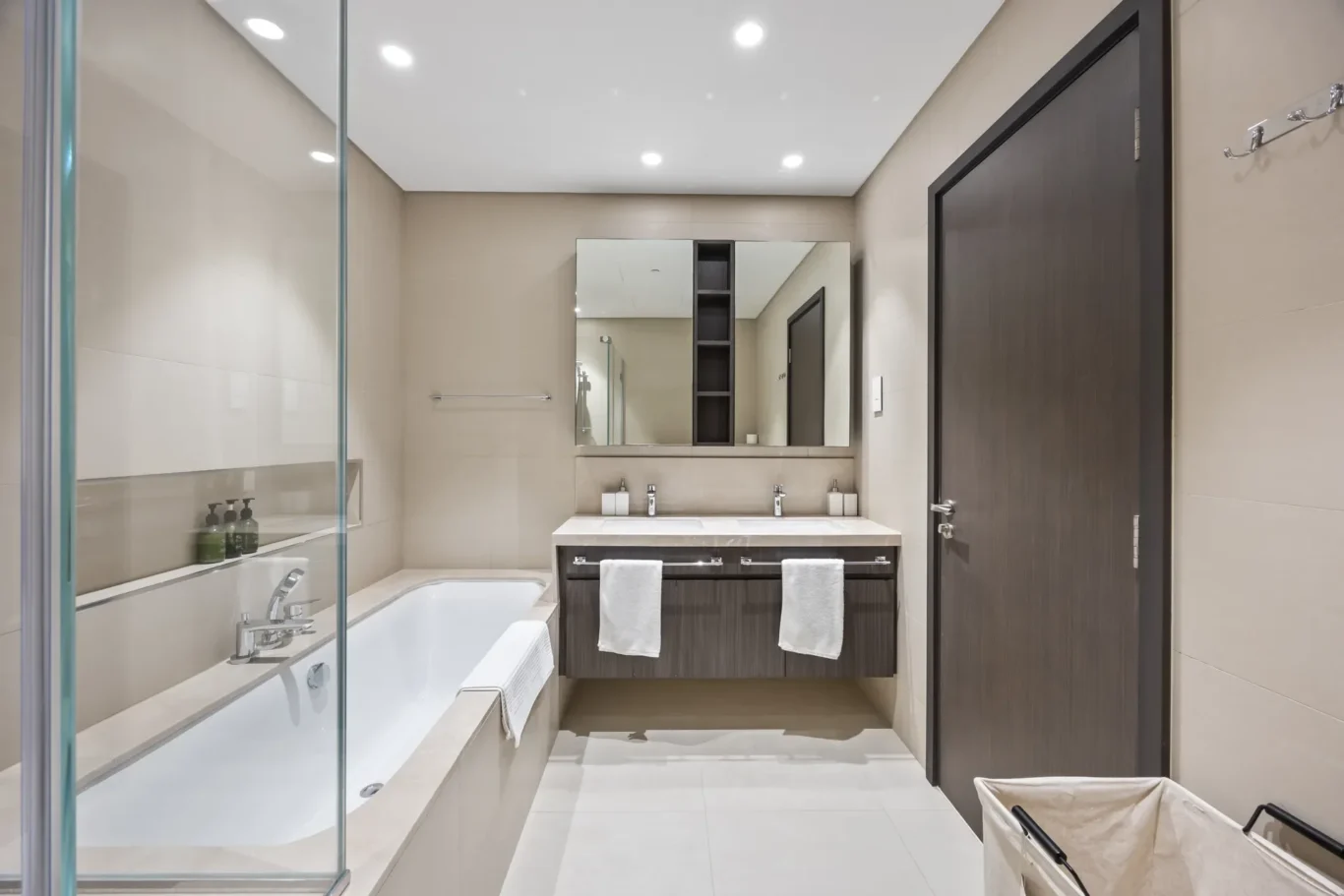 Modern beige-tiled bathroom with a double-sink vanity, large mirror, and glass-walled shower on the left; a white tub along the wall.