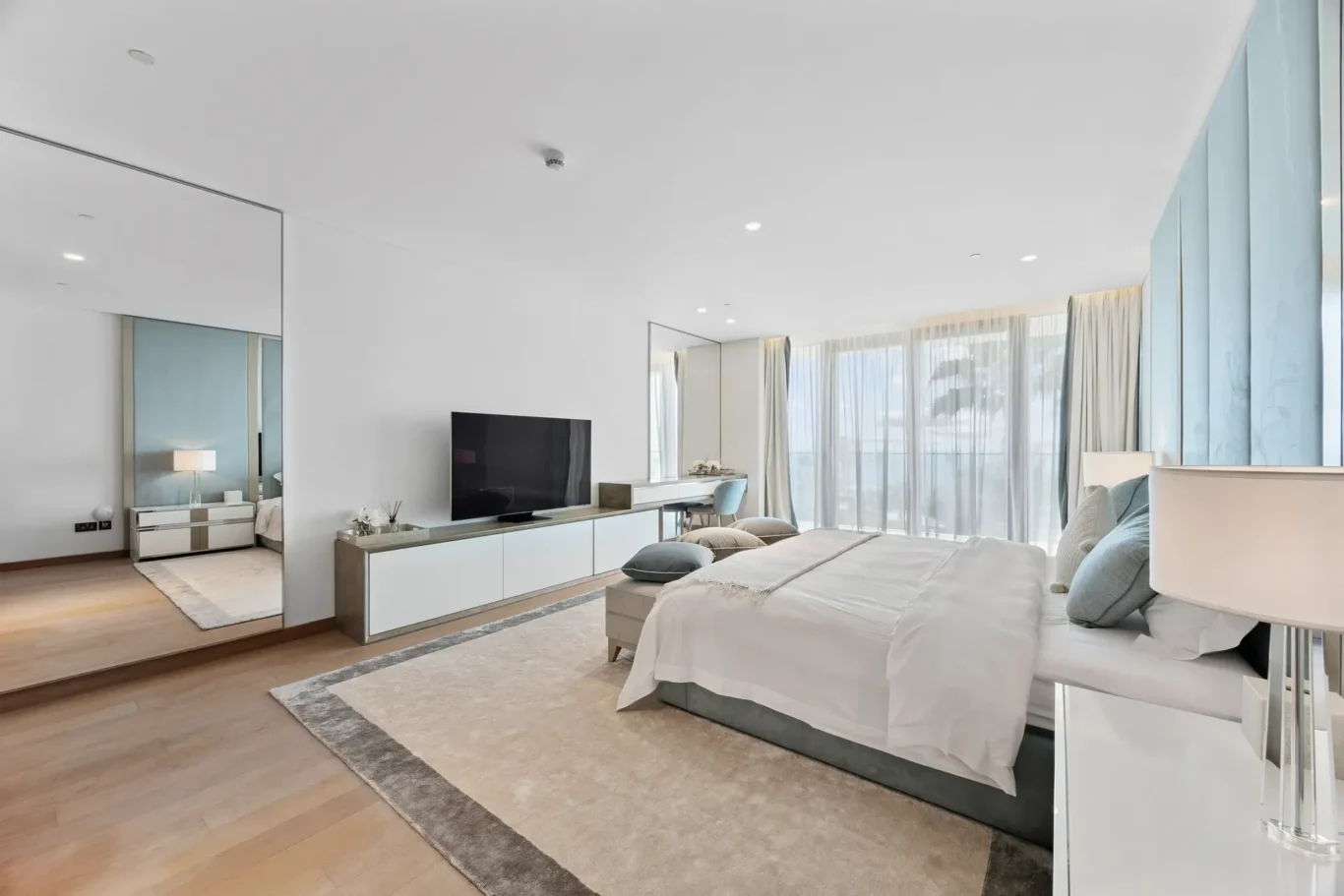 Spacious modern hotel bedroom with a large bed, neutral tones, and floor-to-ceiling windows with sheer curtains. TV on a low console beside the bed.