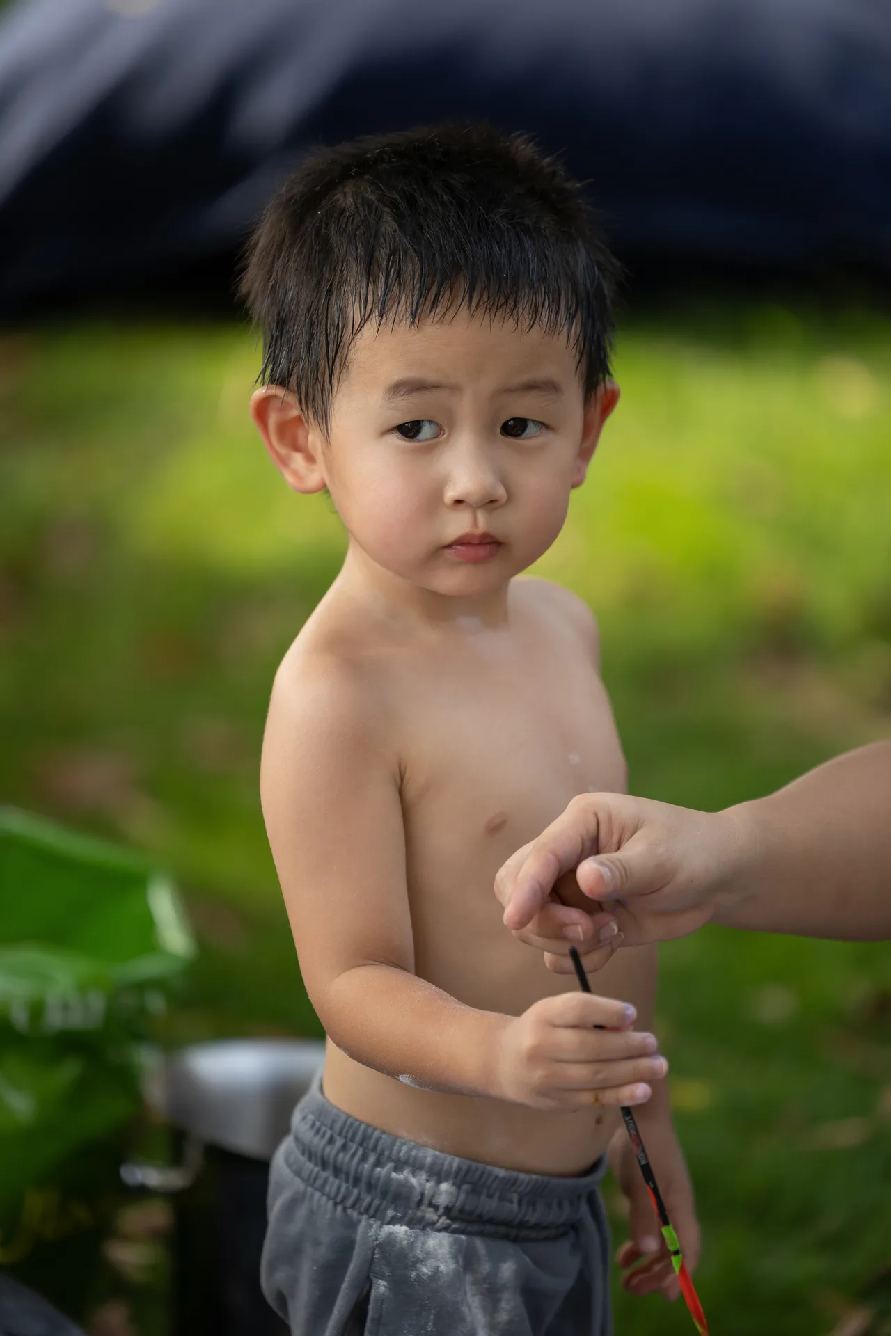 Young child, shirtless, standing outdoors with wet hair, looking to the left as someone holds a string or rope near his hands.