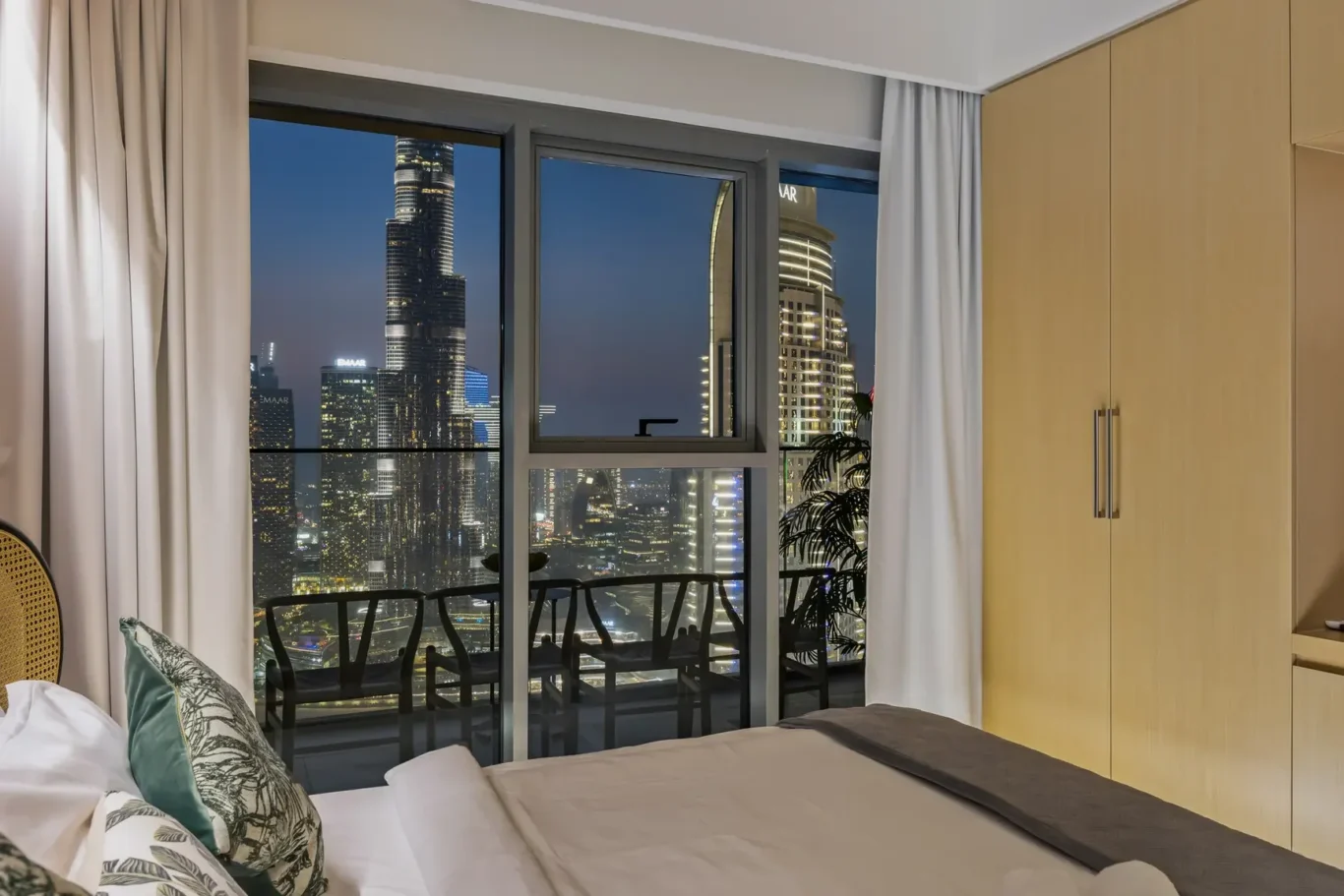 Bedroom with a large window view of a lit city skyline at night; bed with patterned pillows in foreground.