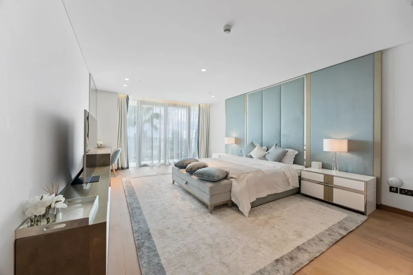 Luxurious hotel bedroom with a king bed, multiple blue pillows, padded headboard panels, and matching nightstands under soft lighting.