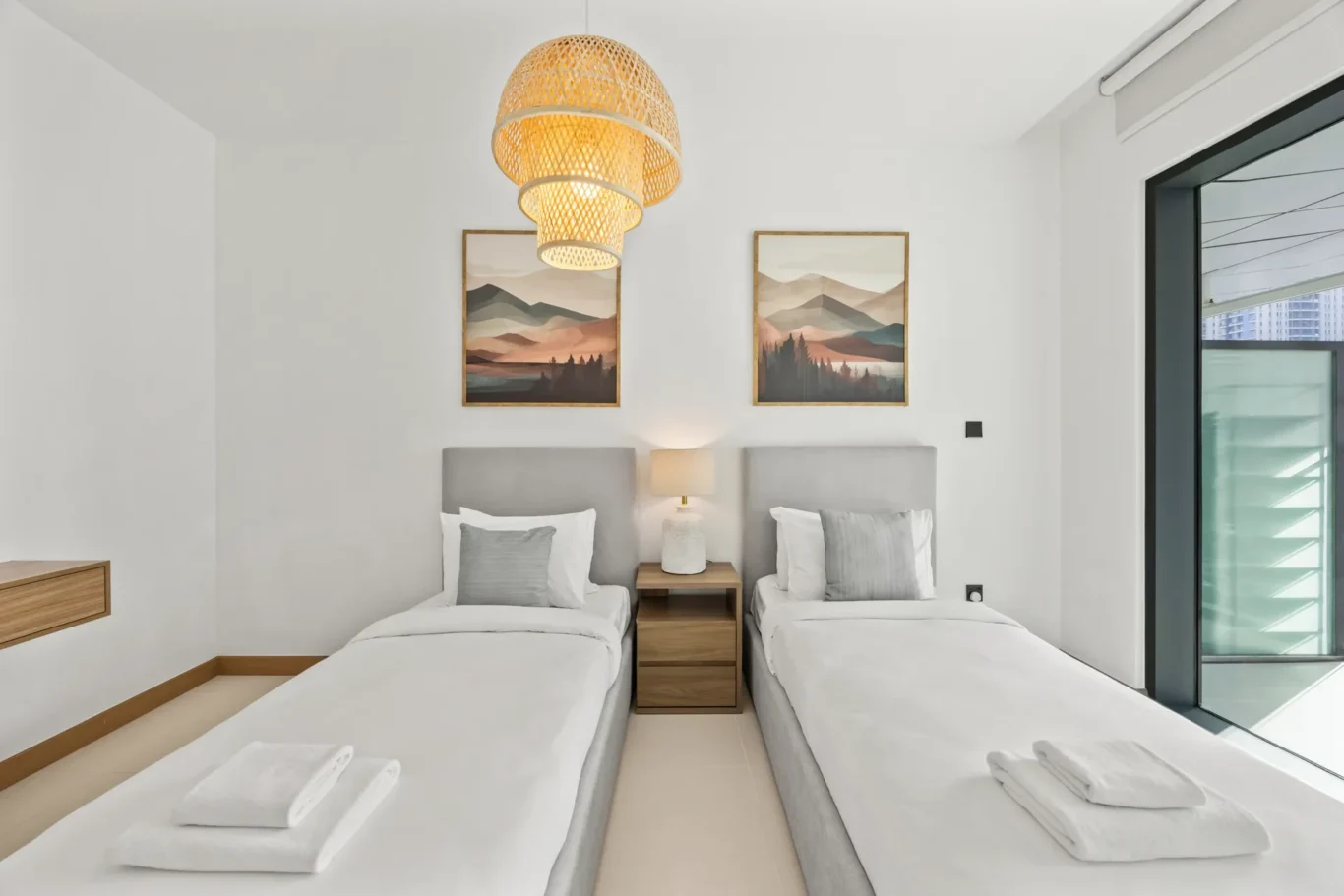 Twin bedroom with two single beds, gray headboards, shared wooden nightstand, and a lamp between them; mountain landscape prints on the wall; bright natural light from a large window.