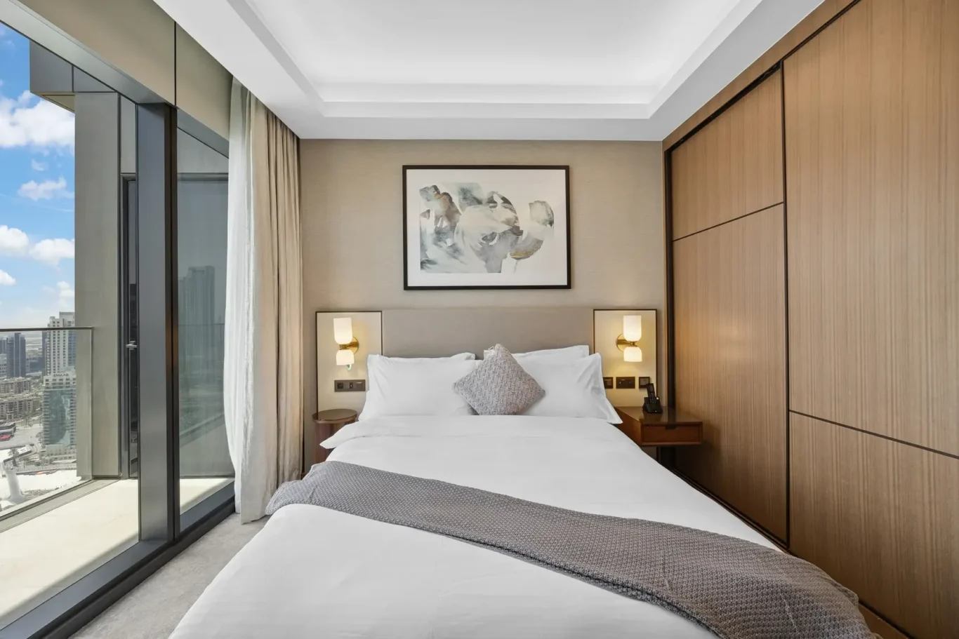 Modern hotel bedroom with a white bed, gray throw, and a framed abstract art piece above the headboard.