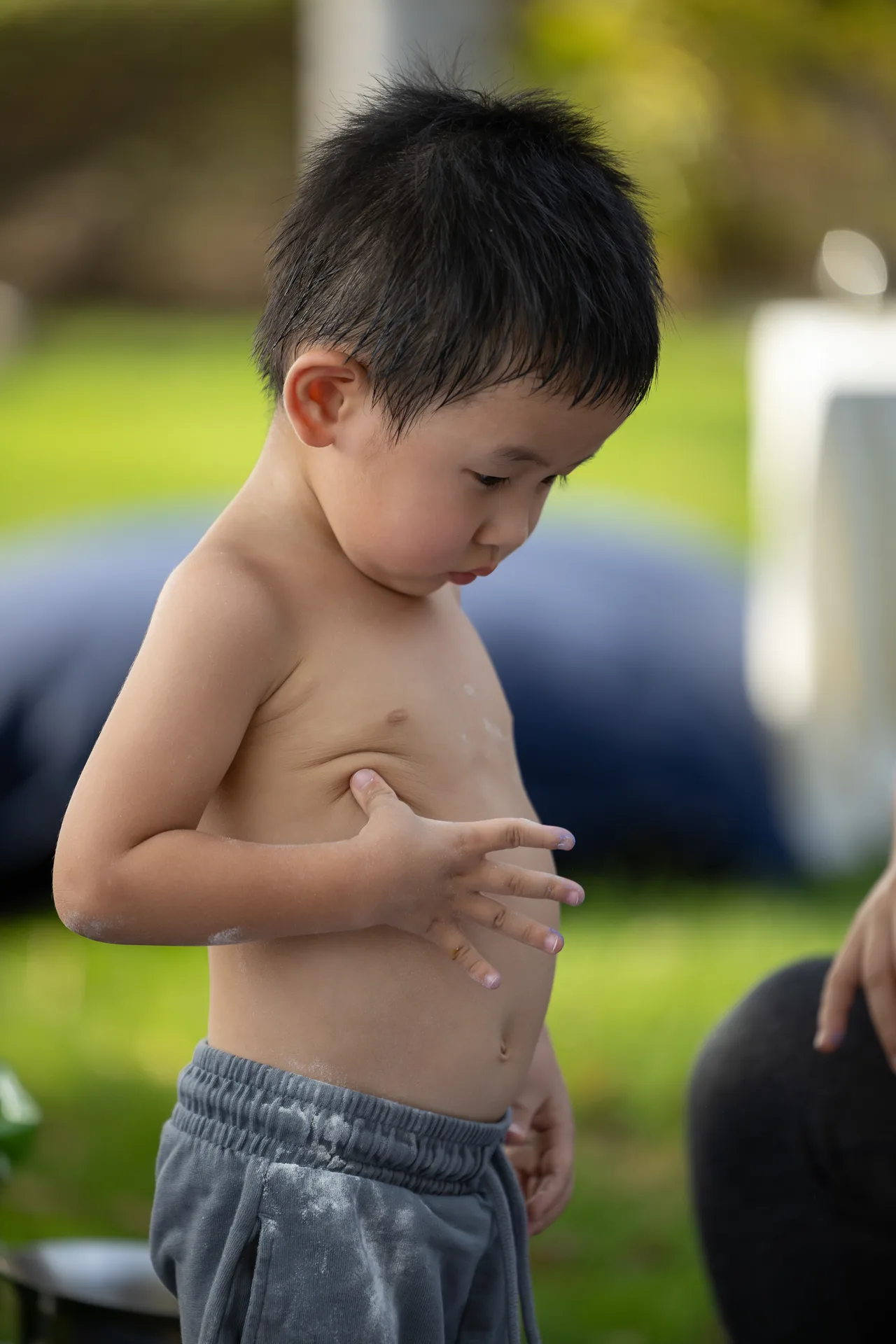 Young boy shirtless outdoors, looking down at his chest with white powder on his torso, wearing gray shorts in a grassy area.