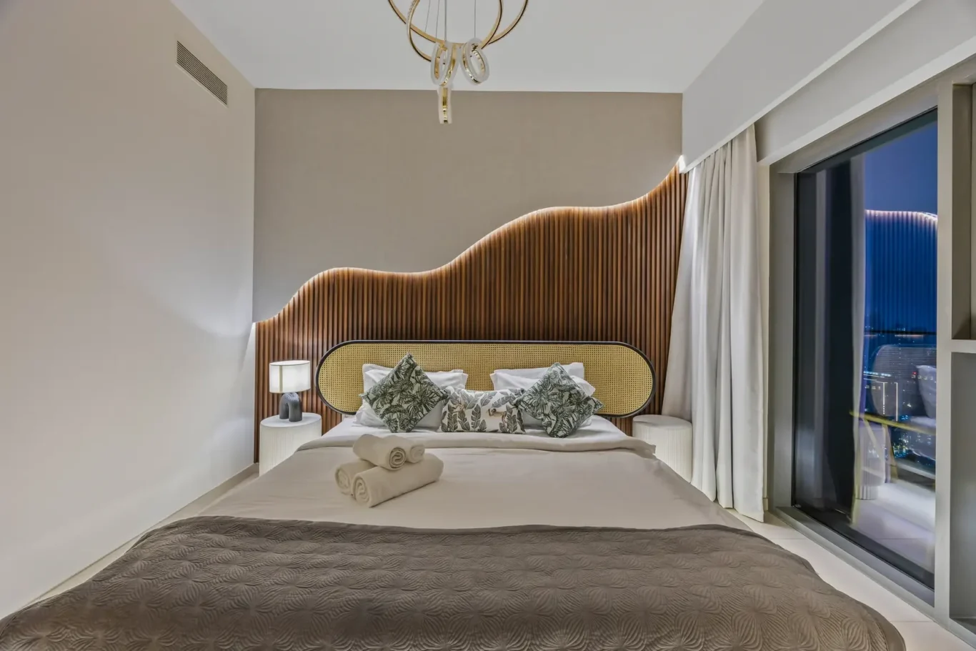 Hotel room bed with curved wooden headboard, botanical pillows, and rolled towels on white bedding; modern nightstands and lamps flank the bed.