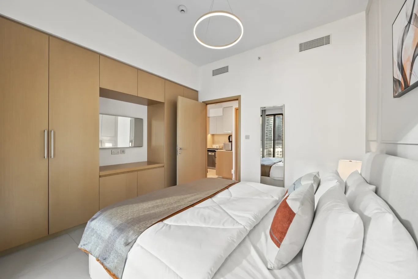 Bright hotel bedroom with a long wooden wardrobe wall, a made double bed with white sheets and orange-gray pillows, and a circular ceiling light.