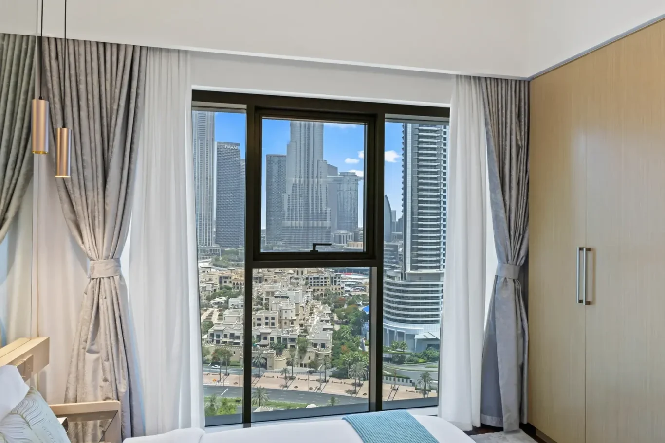 Hotel room with a large window showing the city skyline, gray drapes pulled to the sides, bed in the foreground.",