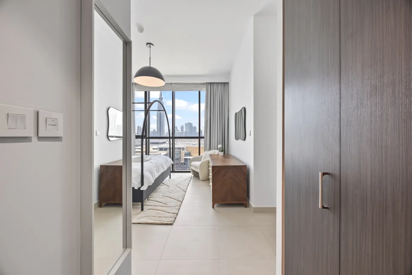 Modern hotel room with a bed facing a large window overlooking the city skyline, plus a desk and mirror.