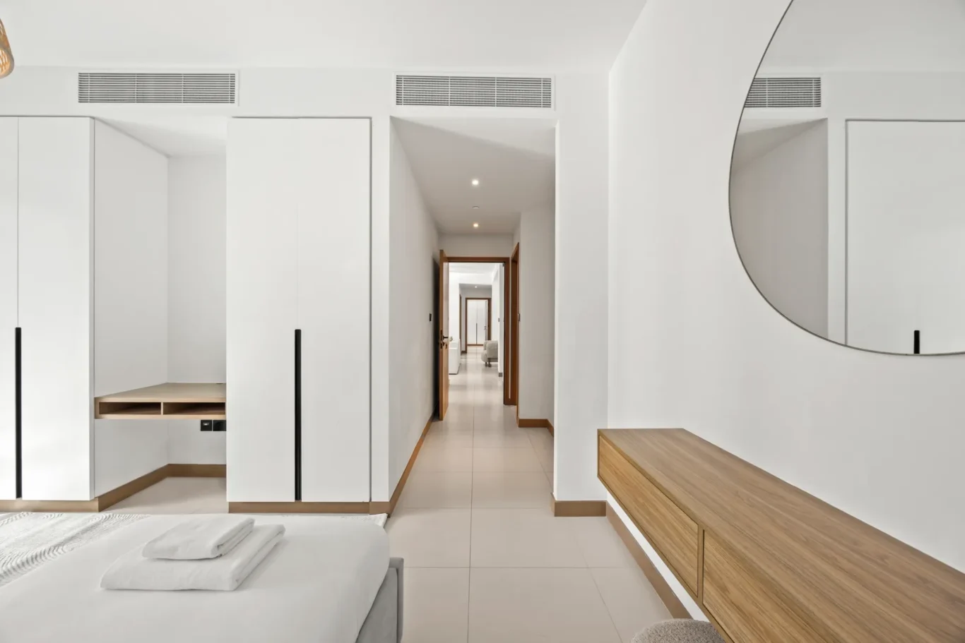 Minimalist hotel bedroom with white built‑in wardrobes, a wooden bench, and a large circular wall mirror.