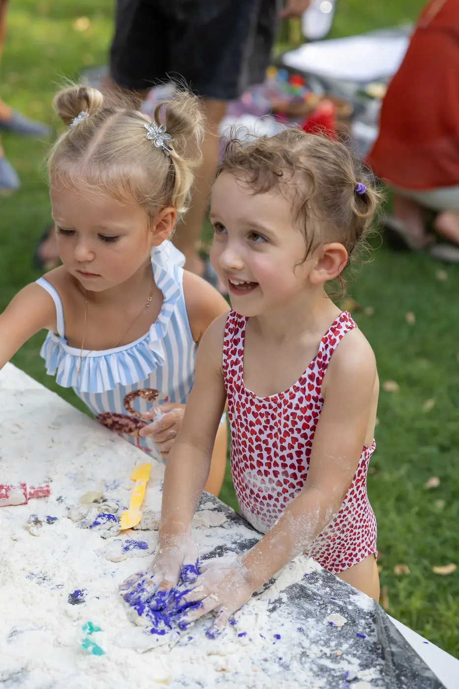 Two young girls in colorful swimsuits play with colored dough at an outdoor table, hands covered in white powder and blue dye.