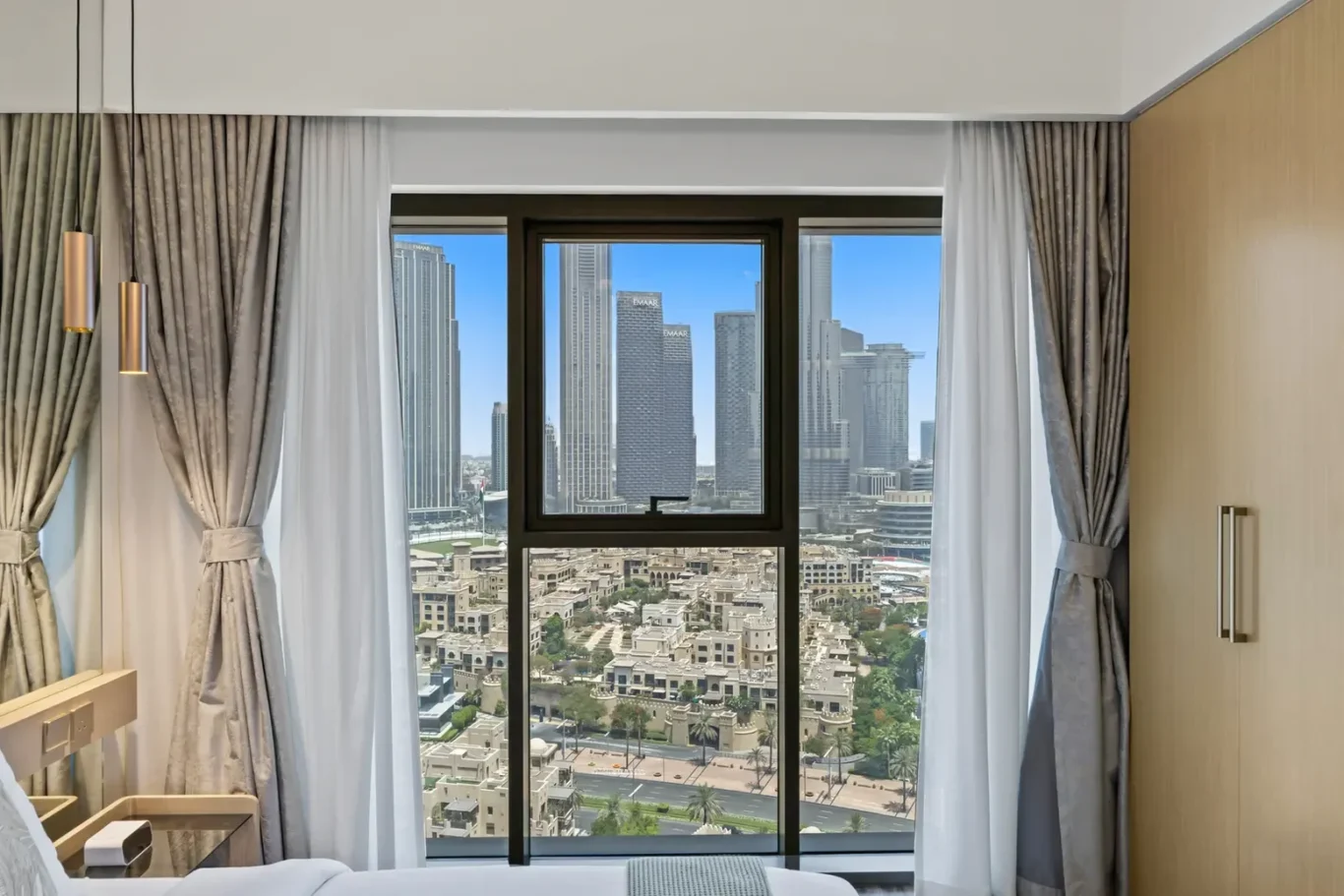 Hotel room with a large window showing a city skyline of tall buildings; sheer white curtains and gray drapes frame the view. The bed and bedside area are visible in the foreground, with a warm interior.