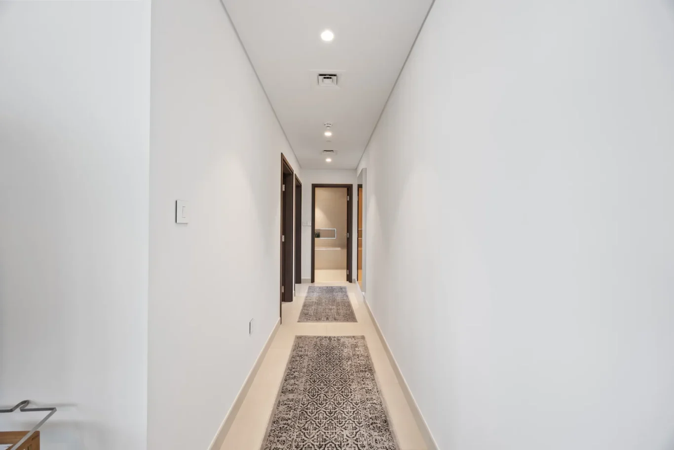 Bright white hallway with recessed ceiling lights and a patterned runner rug leading to a door at the end of the corridor.