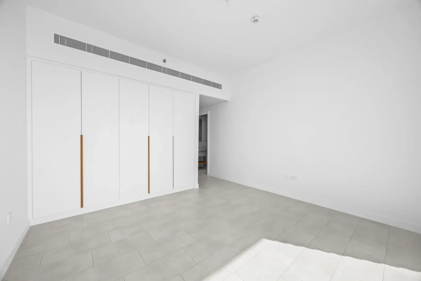 White minimalist room with built-in white wardrobes and vertical gold handles along the left wall, light gray tiled floor