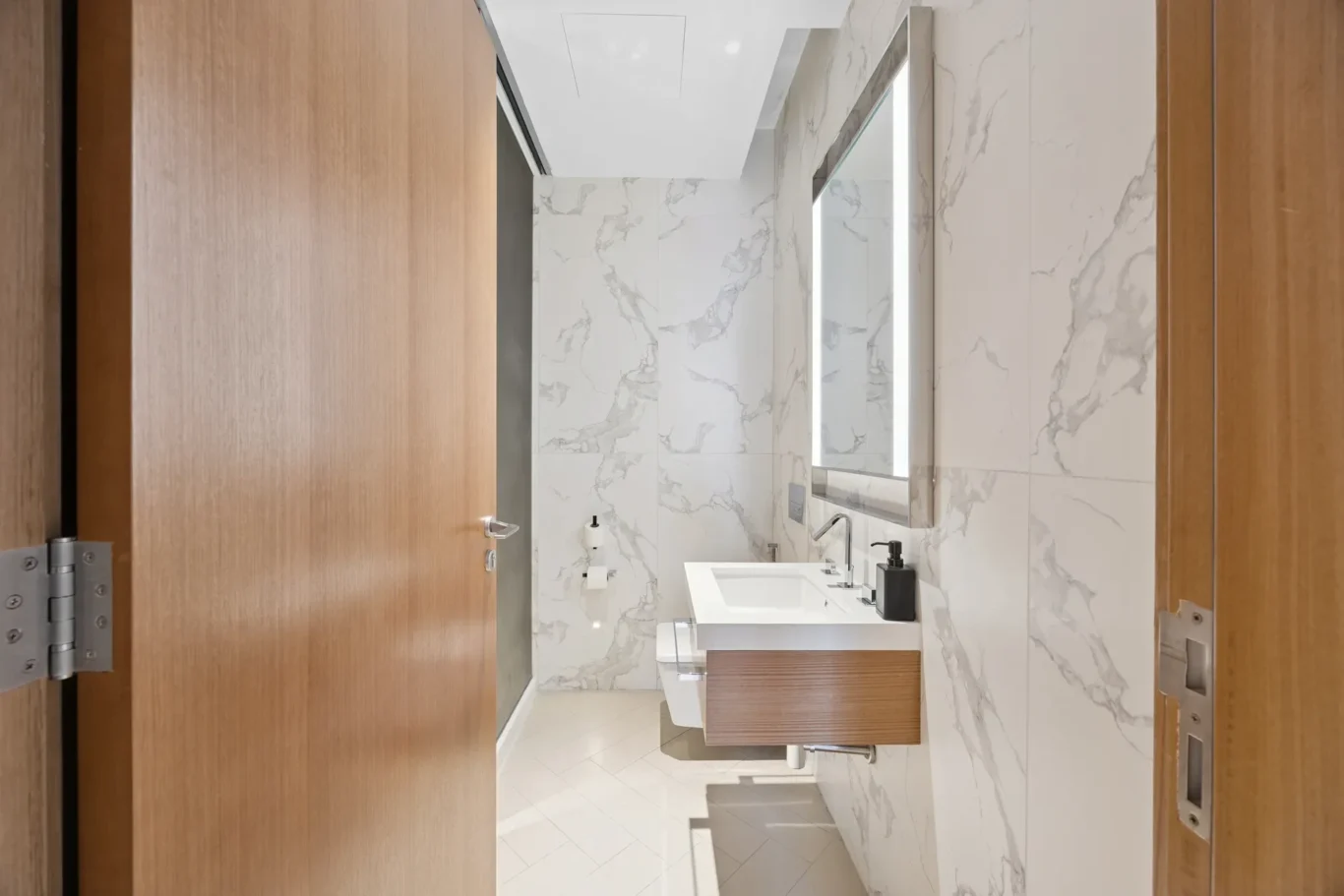 Modern bathroom with marble walls, a backlit rectangular mirror, white sink, and wooden vanity seen from the doorway.