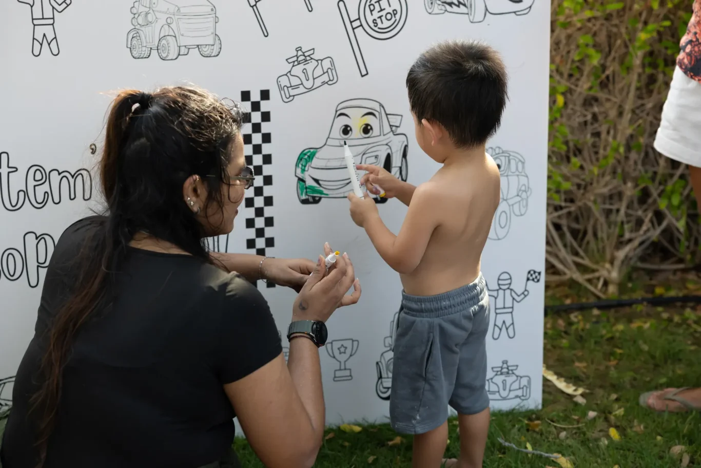 Child coloring a large car-themed board with a white marker while an adult woman helps outdoors.