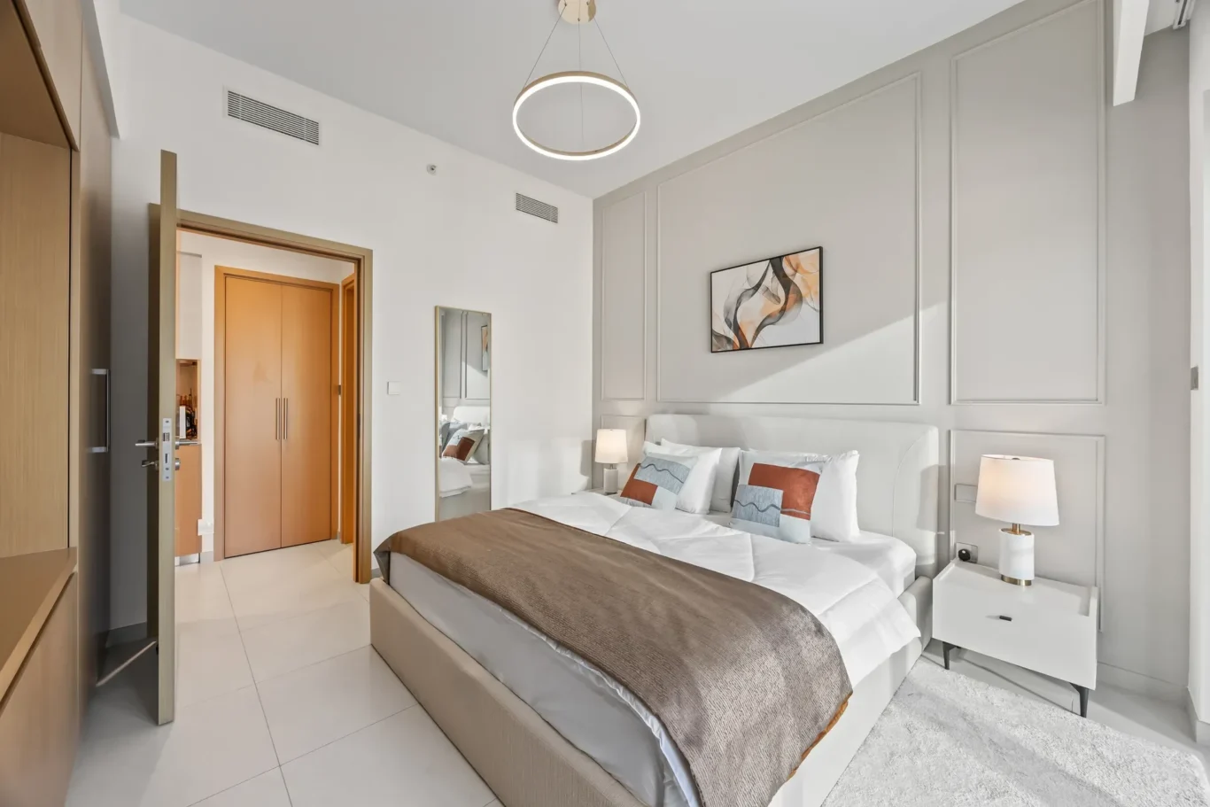 Modern hotel bedroom with a king bed, gray paneled wall, circular ceiling light, abstract wall art, and matching nightstands with lamps.