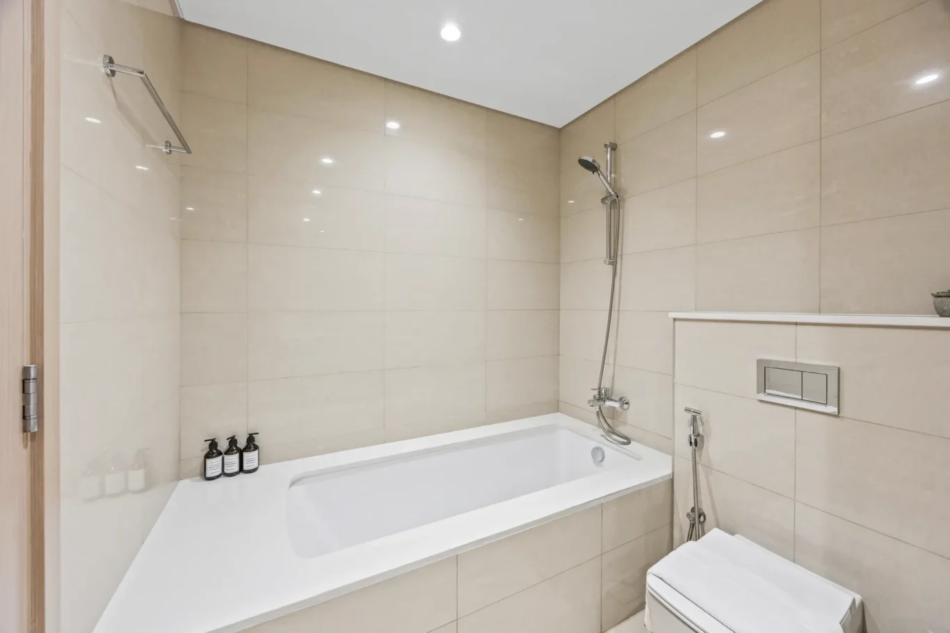 Minimalist beige-tiled bathroom with built-in bathtub, wall-mounted shower and toilet fixtures.