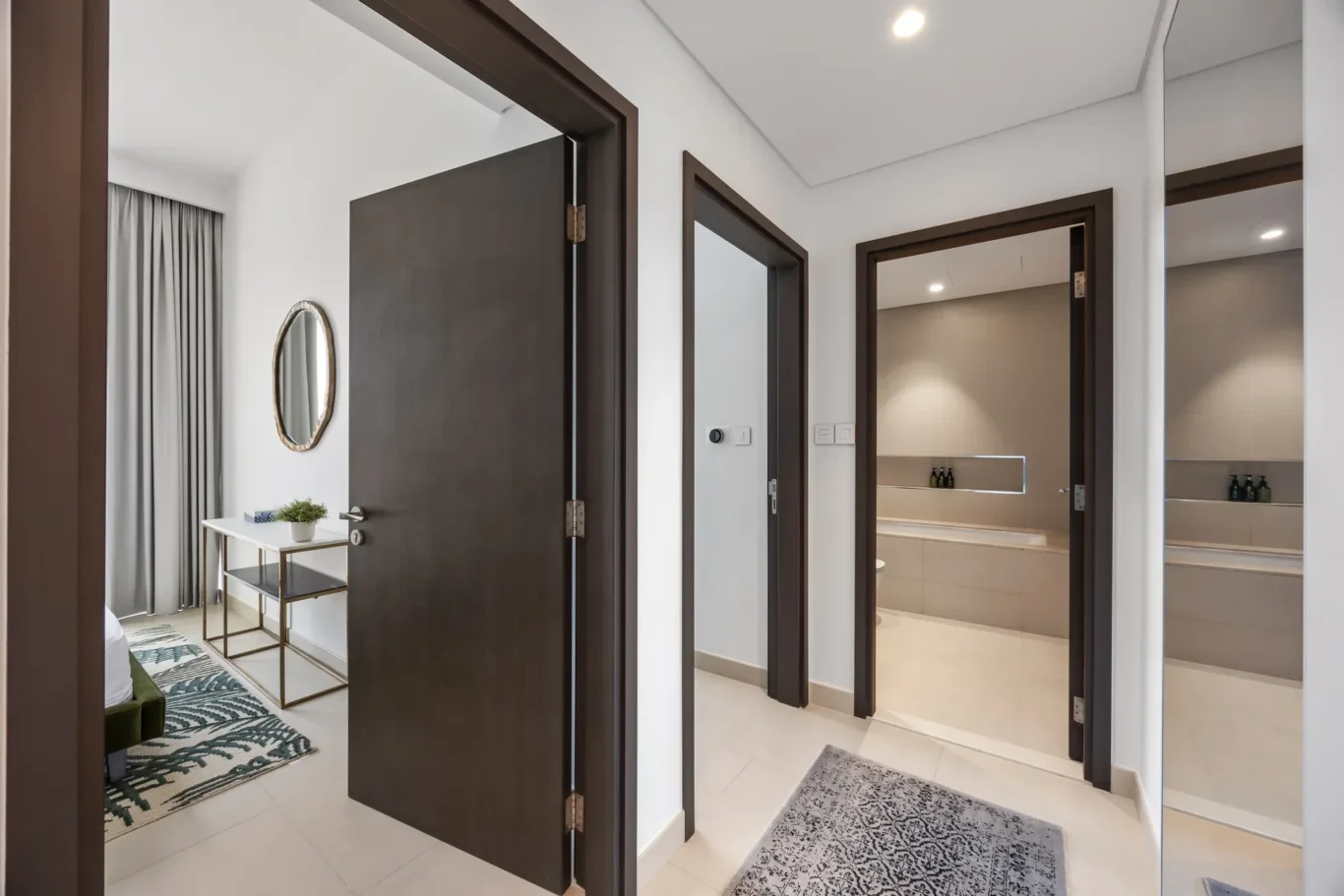Modern hallway with three dark-wood doors; one door partially open reveals a lit bathroom with a vanity and bottles on a shelf in the distance.