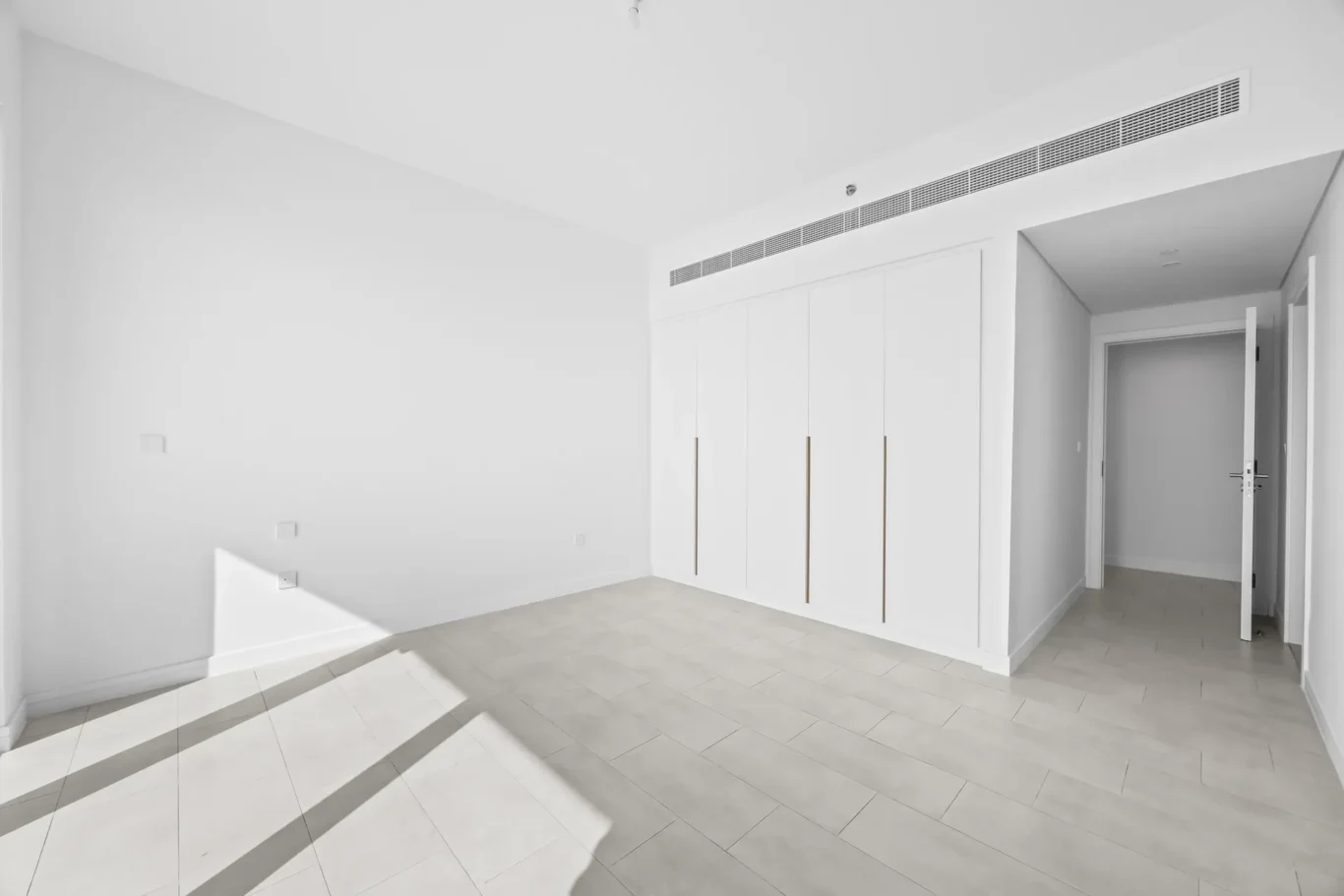 Bright empty room with white built‑in wardrobes along the wall and an open doorway to a hallway.