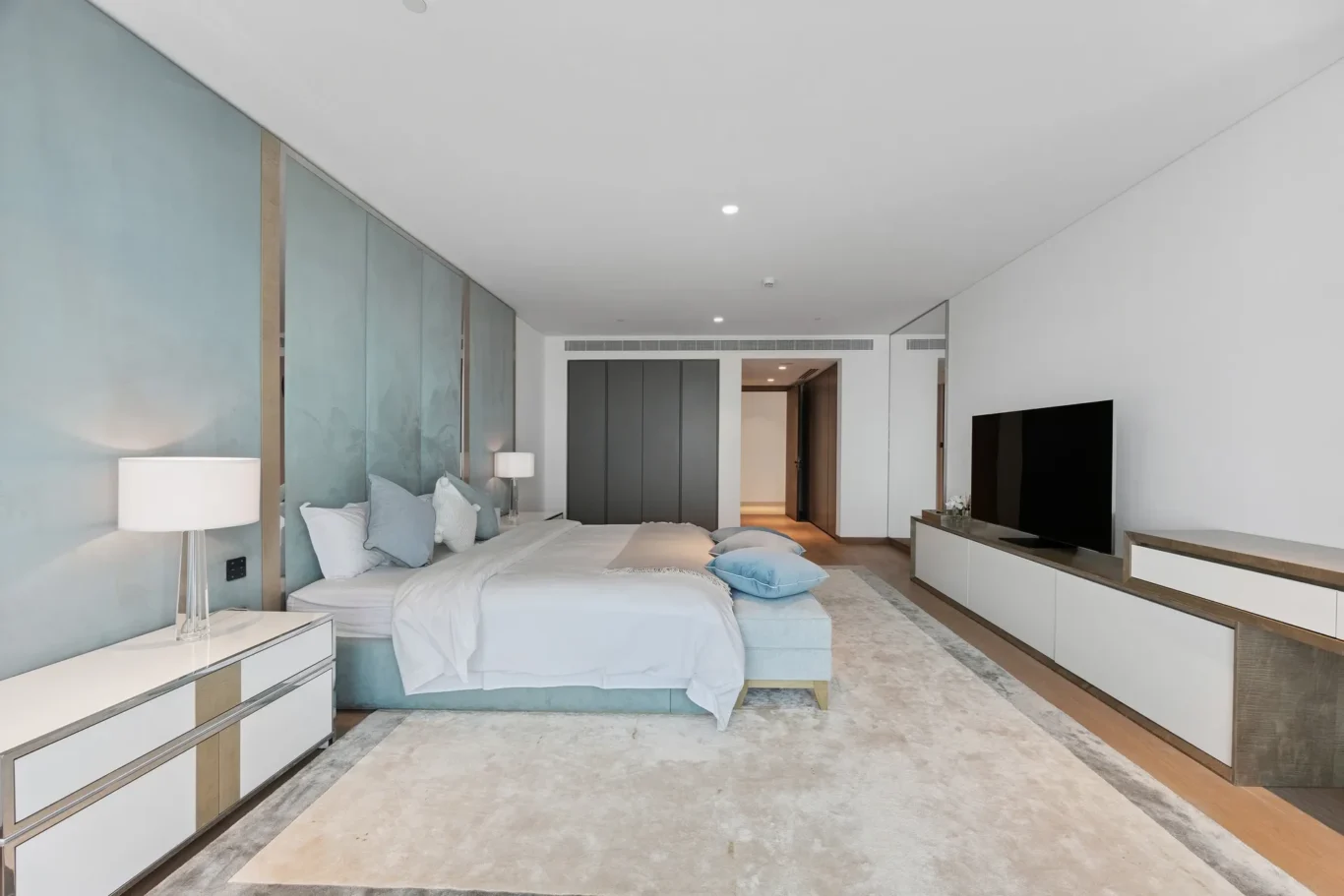 Modern hotel bedroom with a large bed, teal upholstered headboard, and wall-mounted TV opposite.