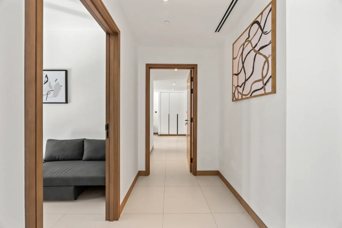 Bright hallway with white walls, wooden door frames, and a grey sofa visible in an adjacent room; abstract art hangs on the right wall.