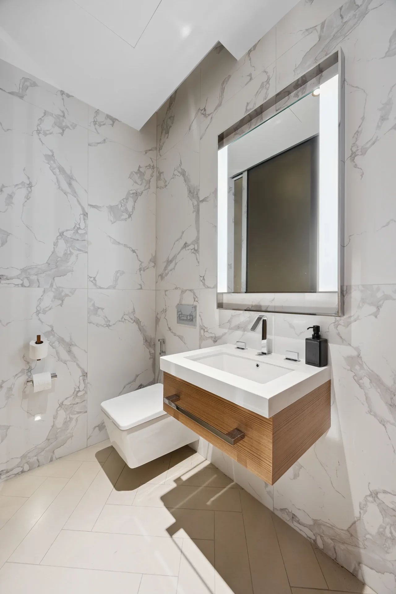 Modern bathroom with marble walls, wall-mounted wooden vanity, rectangular backlit mirror, and wall-hung toilet.