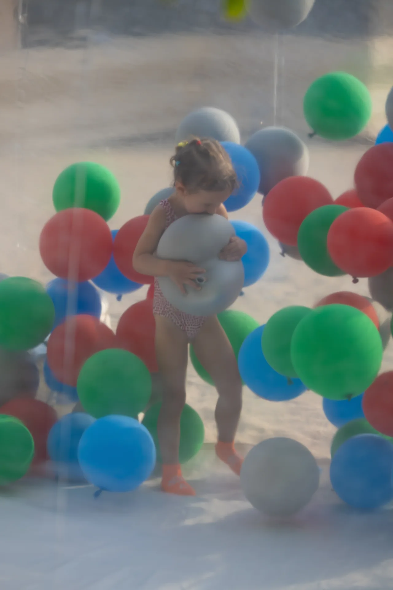 Child wearing a swimsuit hugs a large gray balloon inside a clear bubble surrounded by colorful balloons (red, green, blue) on a sunny day
