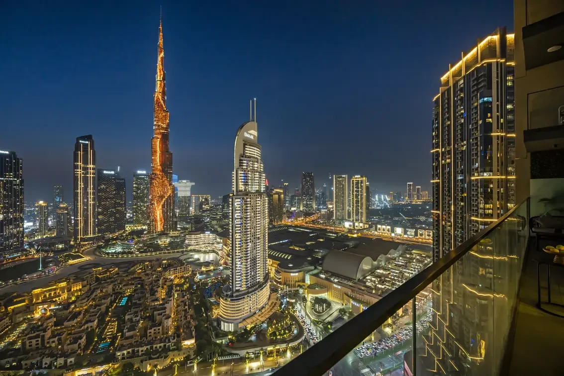 Nighttime Dubai skyline with illuminated skyscrapers, including the Burj Khalifa, seen from a balcony.