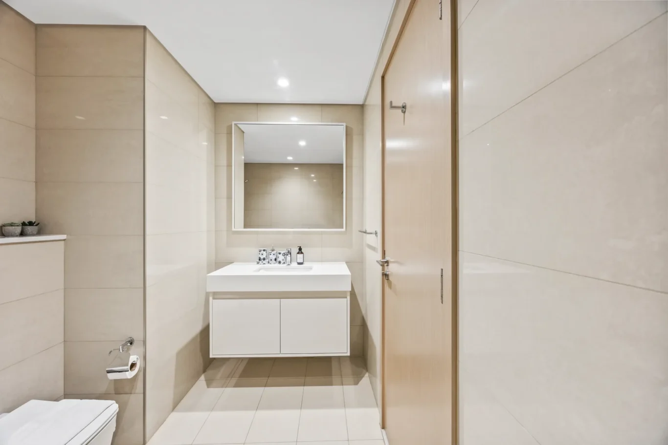 Modern beige-tiled bathroom with a white wall-mounted vanity, square mirror, and a wooden door on the right side; small potted plants on a ledge to the left.