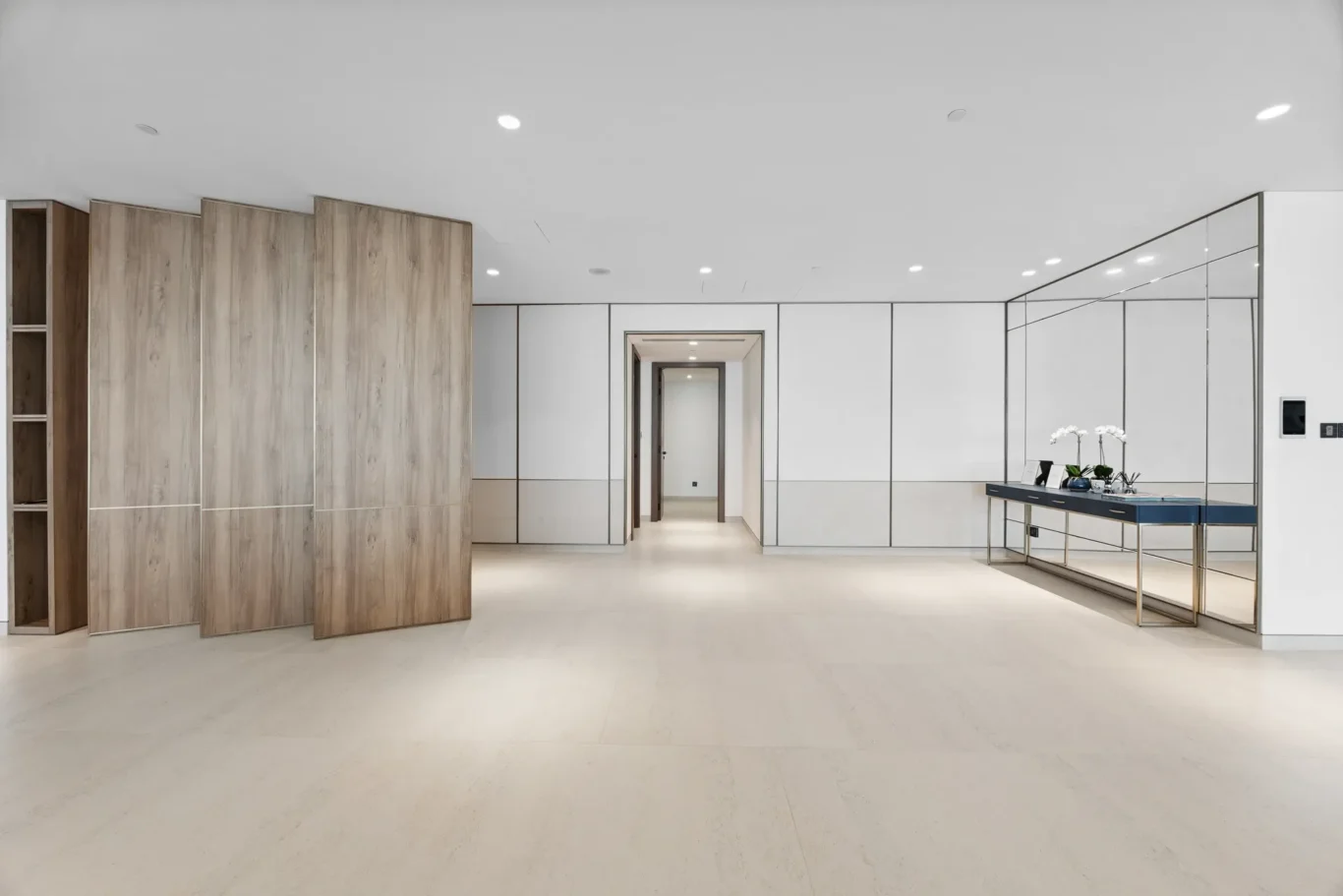 Modern minimalist interior with wood-paneled wall on left, white paneled walls, and a glass mirrored wall on the right featuring a console table with orchids at the far end.