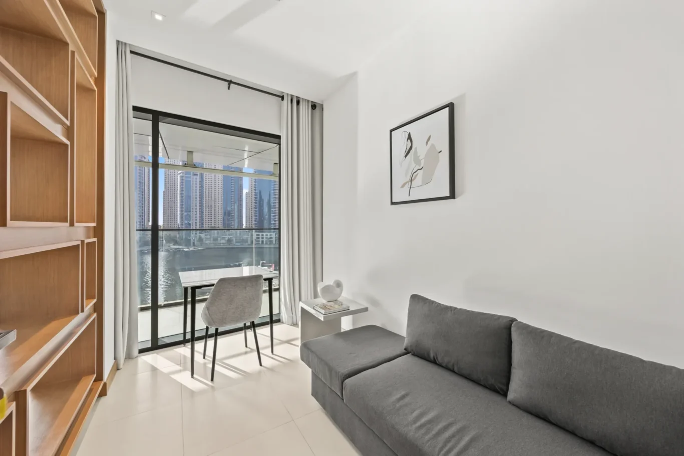 Modern living area with a gray sectional, white walls, abstract framed art, and a balcony with a water-facing city skyline outside.