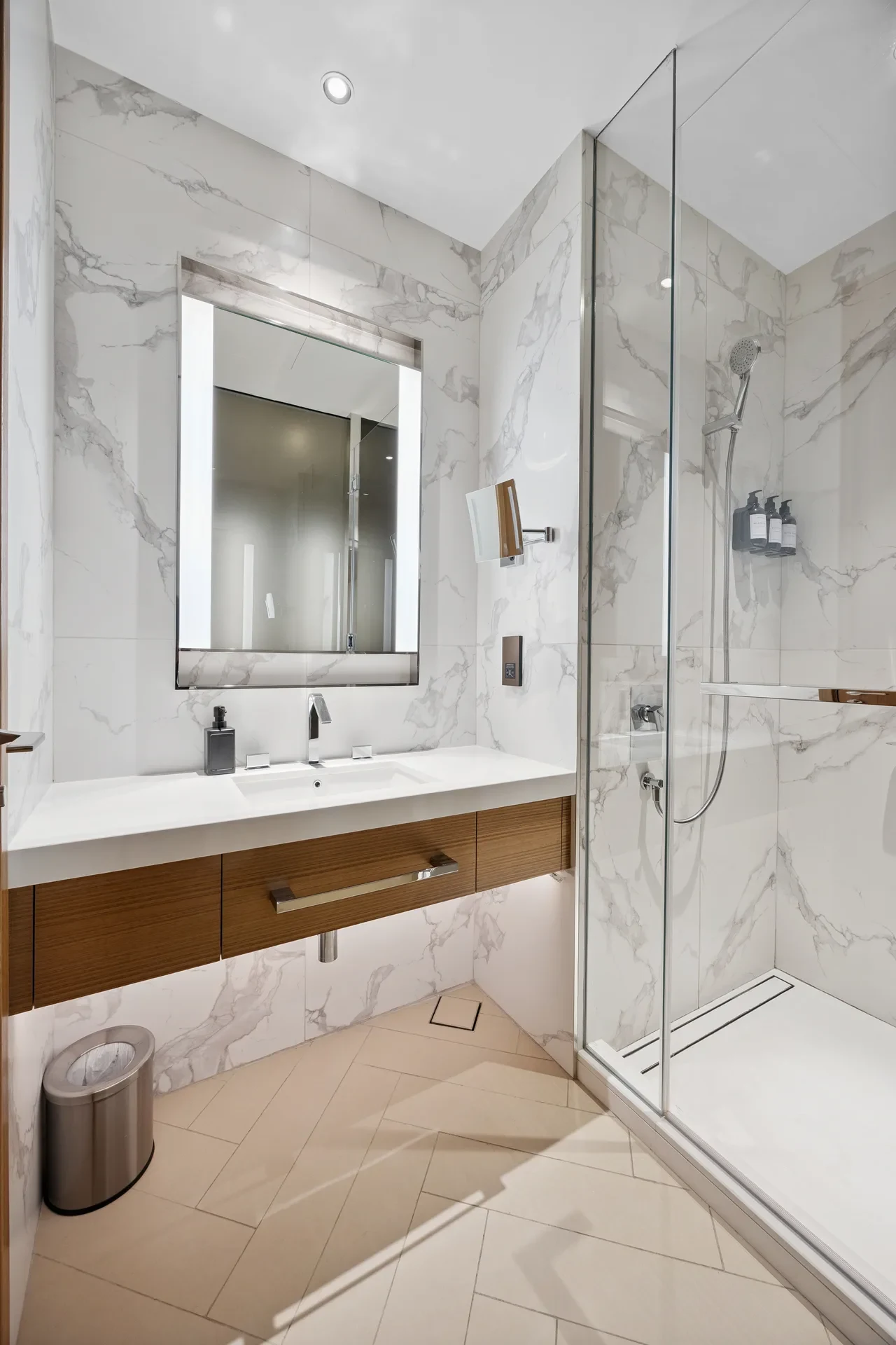 Modern marble-tiled bathroom with a wall-mounted wooden vanity, large backlit mirror, and glass shower enclosure.