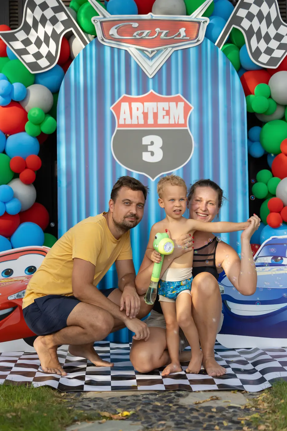 Family posing at a Cars-themed birthday backdrop for Artem's 3rd birthday; child holds a water toy while parents kneel beside him.
