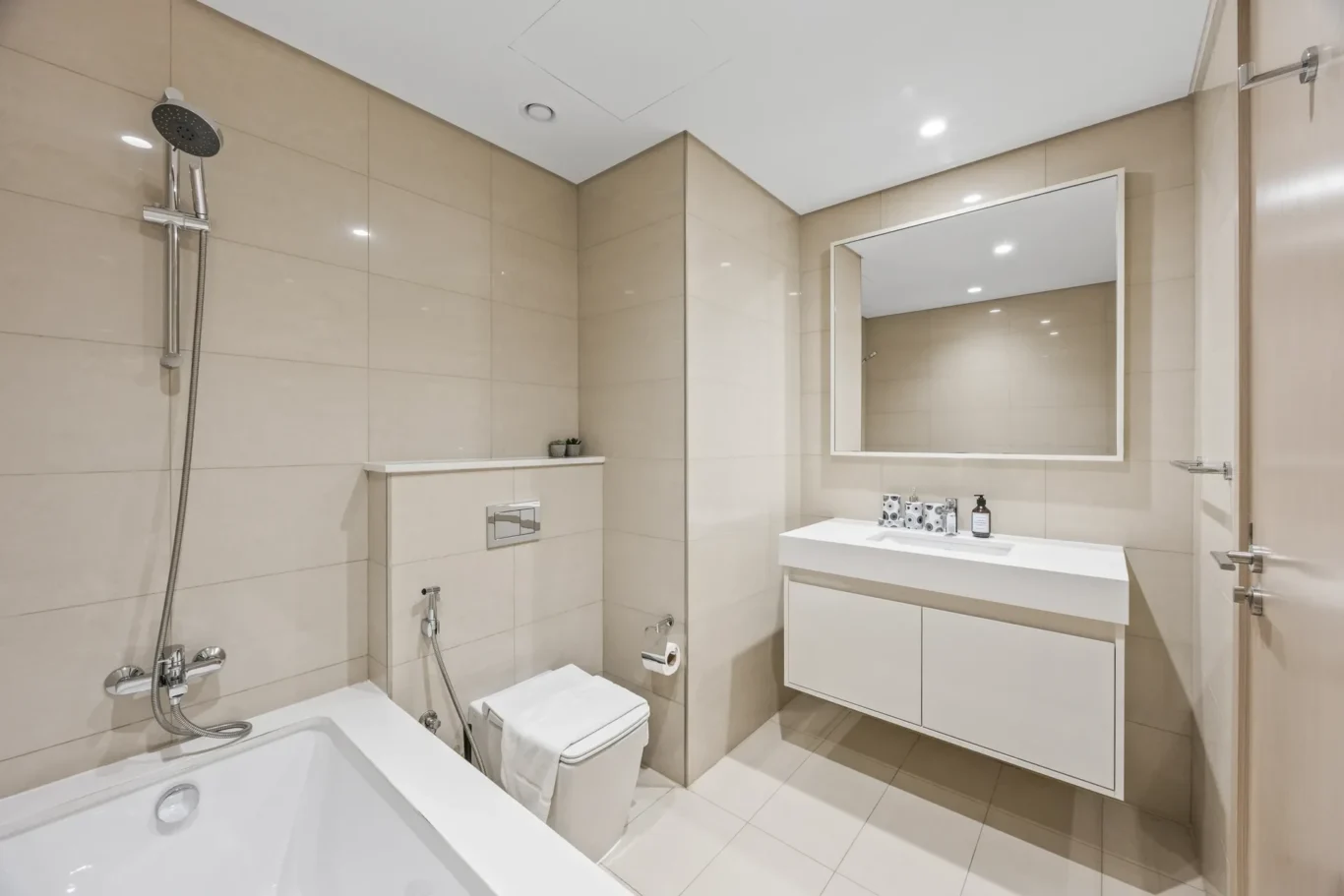 Modern beige-tiled bathroom with a white bathtub, toilet, and floating vanity with a large mirror and chrome fixtures.