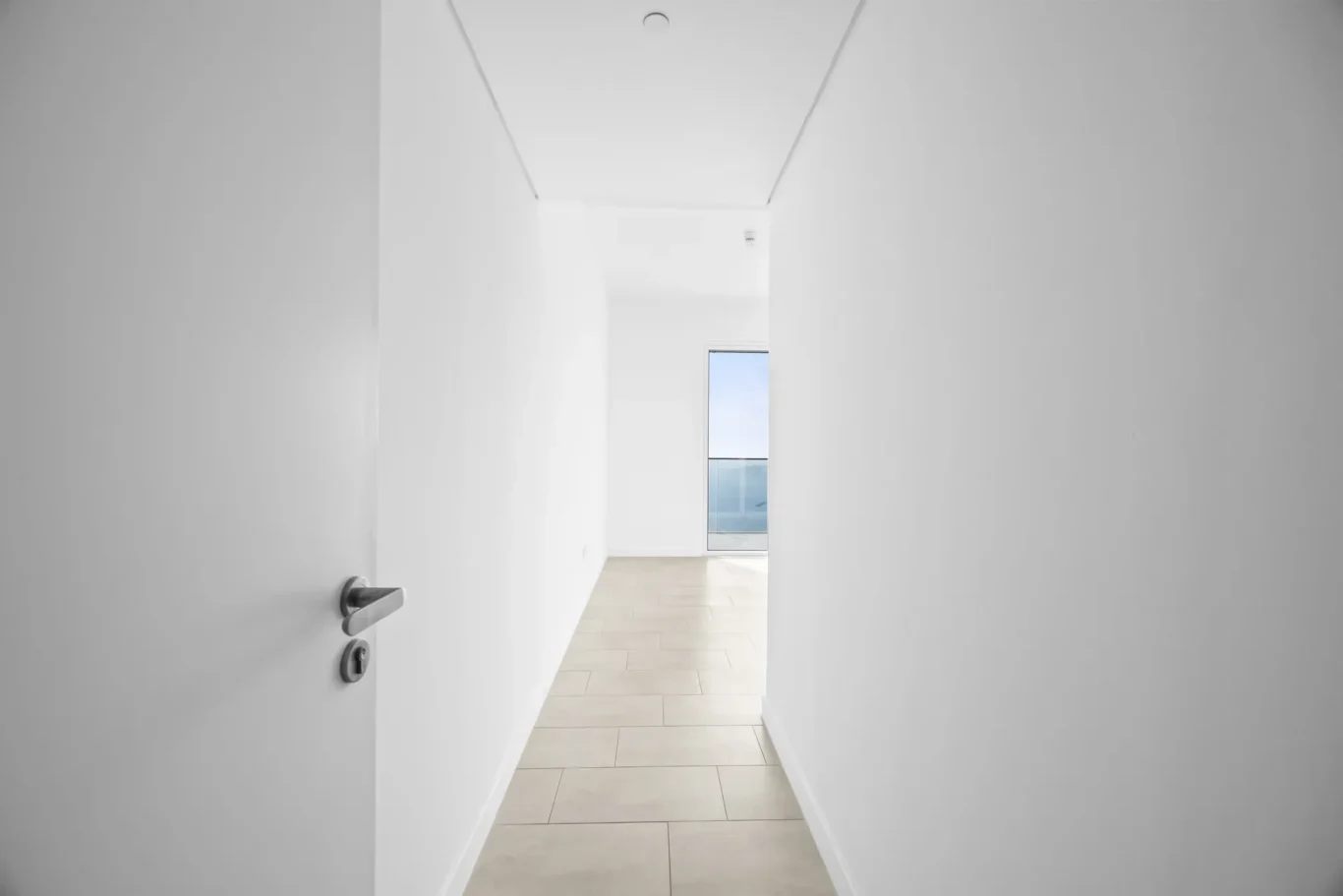 Bright white hallway seen from an open door, leading to a glass door at the far end with blue sky outside