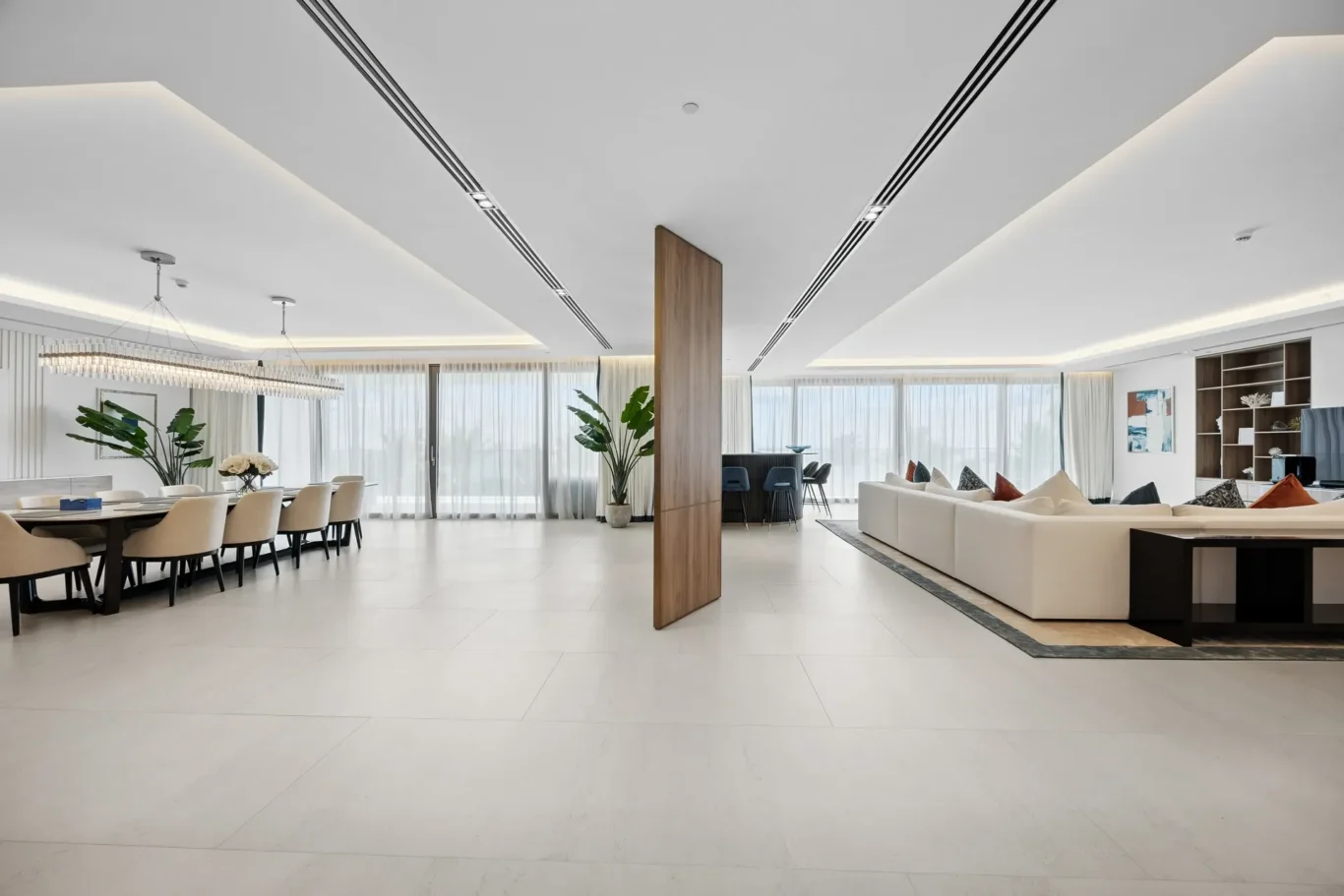 Open-concept living and dining room with a long dining table, beige chairs, and a white sectional sofa by large windows.