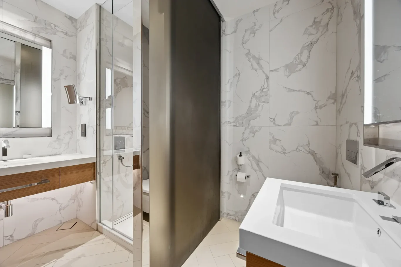 Modern bathroom with white marble walls and a glass-framed shower, wood vanity, and white rectangular sink with a wall-mounted faucet.