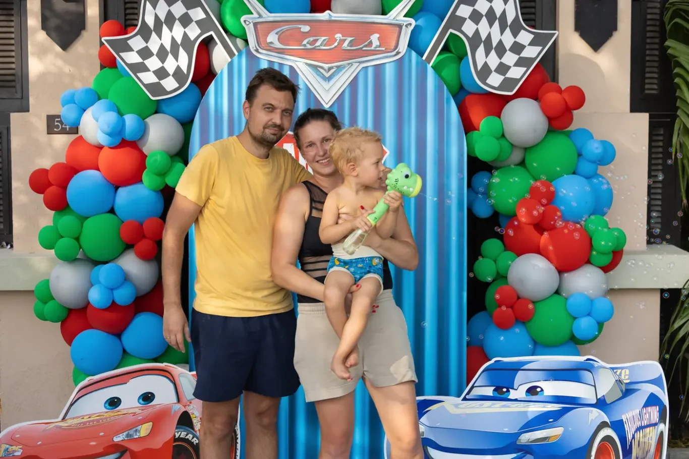 Family of three posing in front of a Cars-themed photo backdrop with colorful balloon arches and cutout cars.