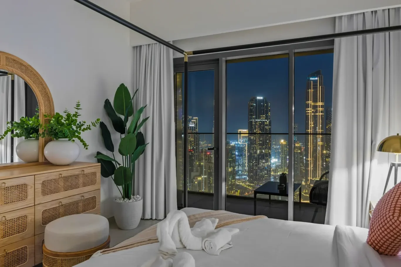 Hotel bedroom with floor-to-ceiling windows showing a lit city skyline at night, white bedding, and a wooden dresser with green plants and a round wicker mirror reflectivity on the wall.