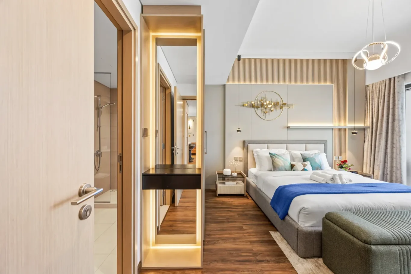 Modern hotel bedroom with a large upholstered bed, white bedding, blue throw, and gold circular wall art above the headboard; warm wooden slat wall and backlit mirror feature.