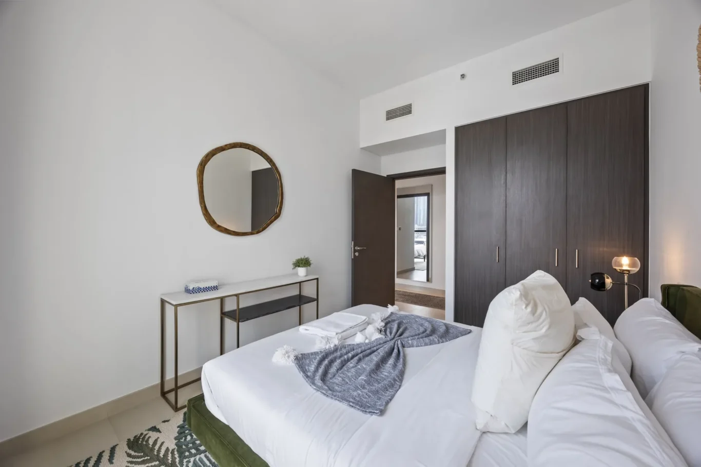 Bright hotel-style bedroom with white bedding, a tall dark wooden wardrobe, and a round gold-framed mirror on the wall.