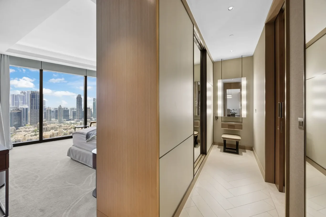 Modern hotel suite hallway with a wooden wardrobe and mirrored panels, leading to a bedroom and a city skyline view outside the window.