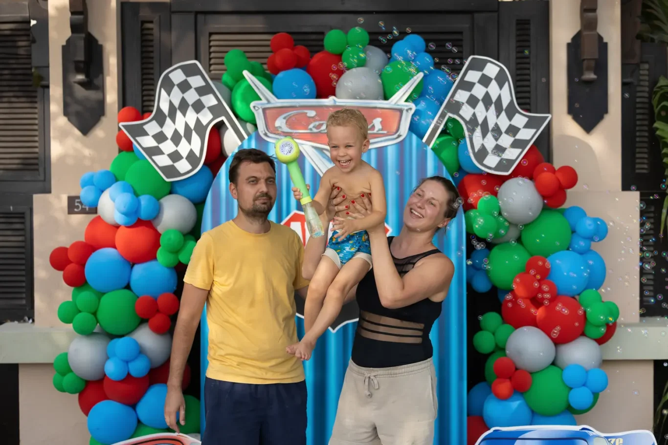 Family of three posing with their toddler at a colorful carnival backdrop of balloons and a car-themed slide, smiling for the camera.