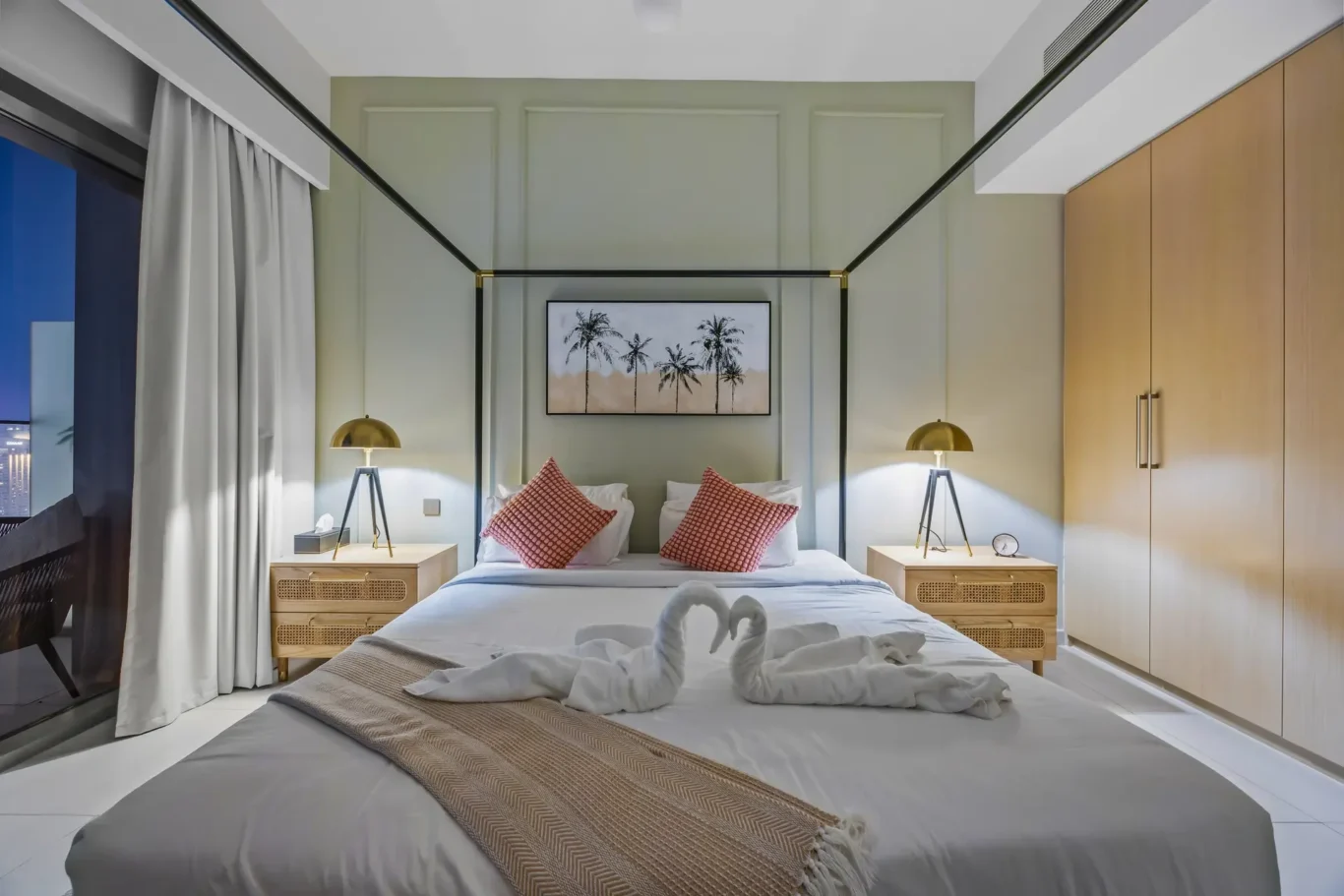 Hotel bedroom with a four-poster canopy bed, palm-framed artwork, and two bedside tables with brass lamps; towels folded into swans on the bed.