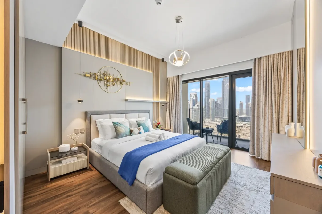 Modern hotel bedroom with a king bed, blue throw, and green ottoman bench facing a balcony with cityscape views.