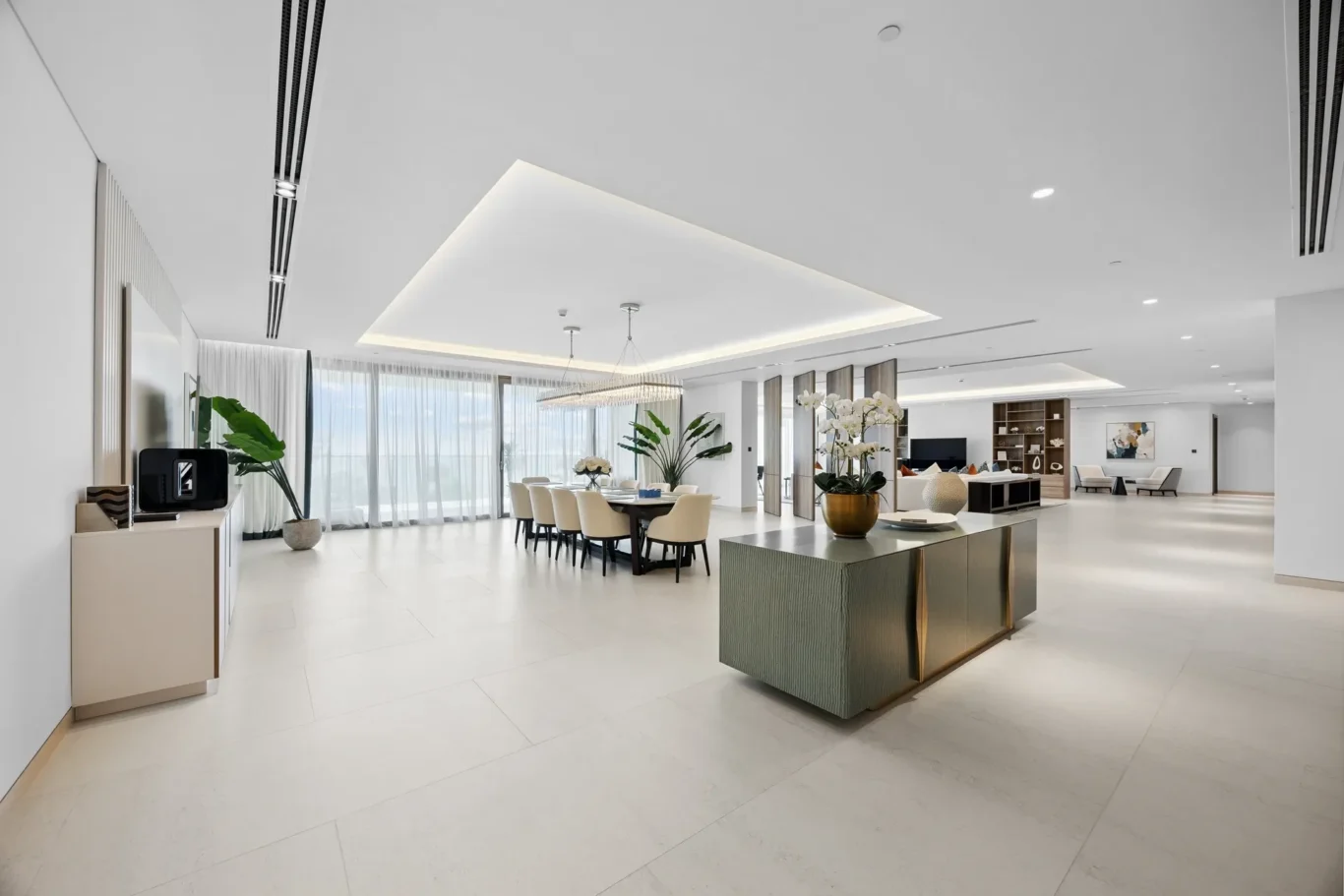 Bright, open-concept living and dining area with a long dining table, beige chairs, chandelier, and large windows with sheer curtains.